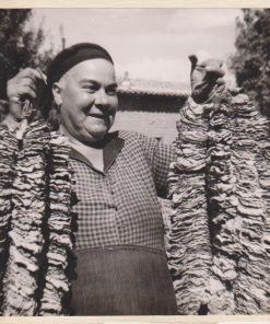 [Καπνά, φωτοκάρτα]: [Γυναίκα κρατεί και επιδεικνύει δύο αρμαθιές καπνών], ασπρόμαυρη επικολλημένη σε χαρτί, διαστάσεις  16.5Χ18