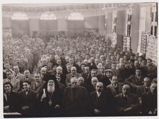 [Σέρρες]: Φωτογραφία, στο πίσω μέρος σημειώνεται με μολύβι: "Από το Συνέδριο Δήμων και Κοινοτήτων Ν. Σερρών. Εις την πρώτη σειρά ο Σεβασμιώτατος Σιδηροκάστρου...οι βουλευταί Σερρών κ. Μόντζαλας και Μηταφίδης / 14,15 και 16 Μαρτίου 1955", ασπρόμαυρη, διαστάσεις 18Χ12,  τσάκισμα στο επάνω μέρος